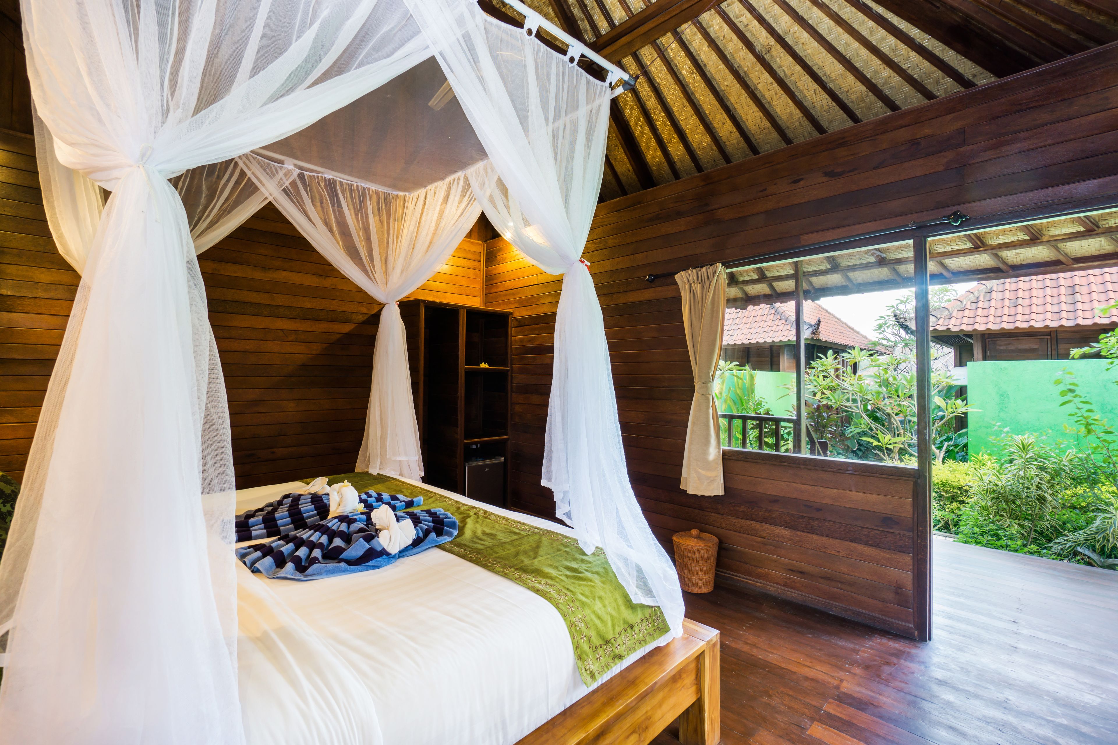 A wooden room with a bed under a white canopy, a window with a view of greenery, and a wooden bench.