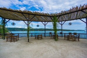 An outdoor seating area with wooden chairs and tables, a thatched roof, and a view of a body of water. - Laguna Reef Huts (Lembongan Island)
