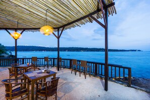 An outdoor seating area with wooden tables and chairs, a thatched roof, and a view of the sea. - Laguna Reef Huts (Lembongan Island)