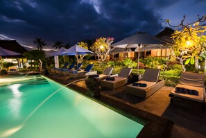A nighttime pool area with lounge chairs and umbrellas, surrounded by lush greenery and a mountain backdrop. - Laguna Reef Huts (Lembongan Island)