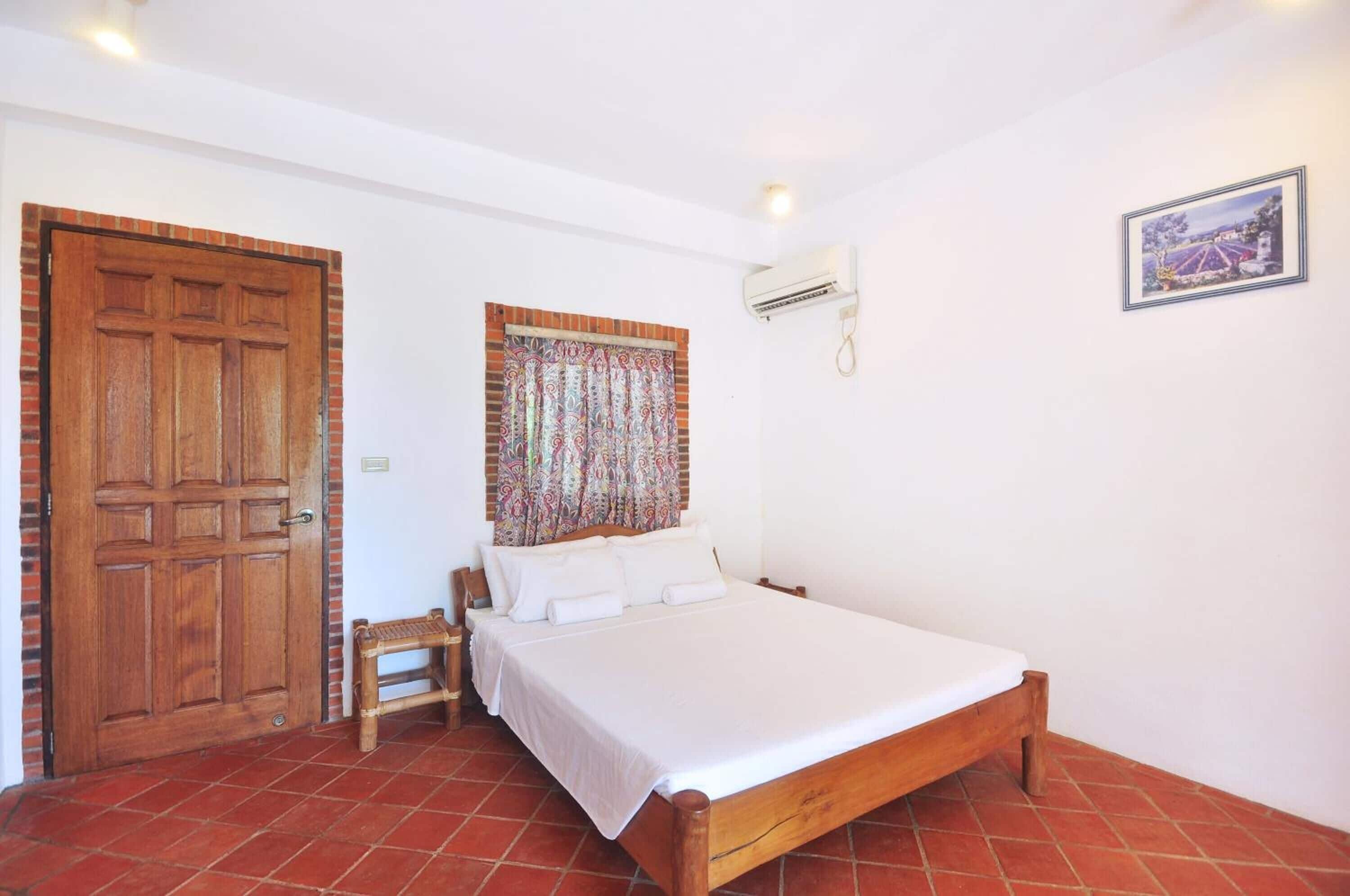 A bedroom with a wooden bed, a small wooden stool, a patterned curtain, a white wall, a framed picture, and a red-tiled floor.