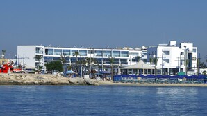 A beachfront hotel with multiple floors, a beach with blue umbrellas, and palm trees. - Limanaki Beach Hotel (Ayia Napa)