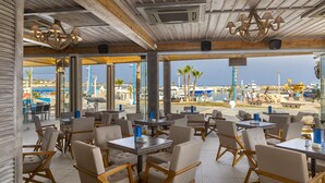 A restaurant with outdoor seating, wooden tables, and chairs, overlooking a beach and marina. - Limanaki Beach Hotel (Ayia Napa)