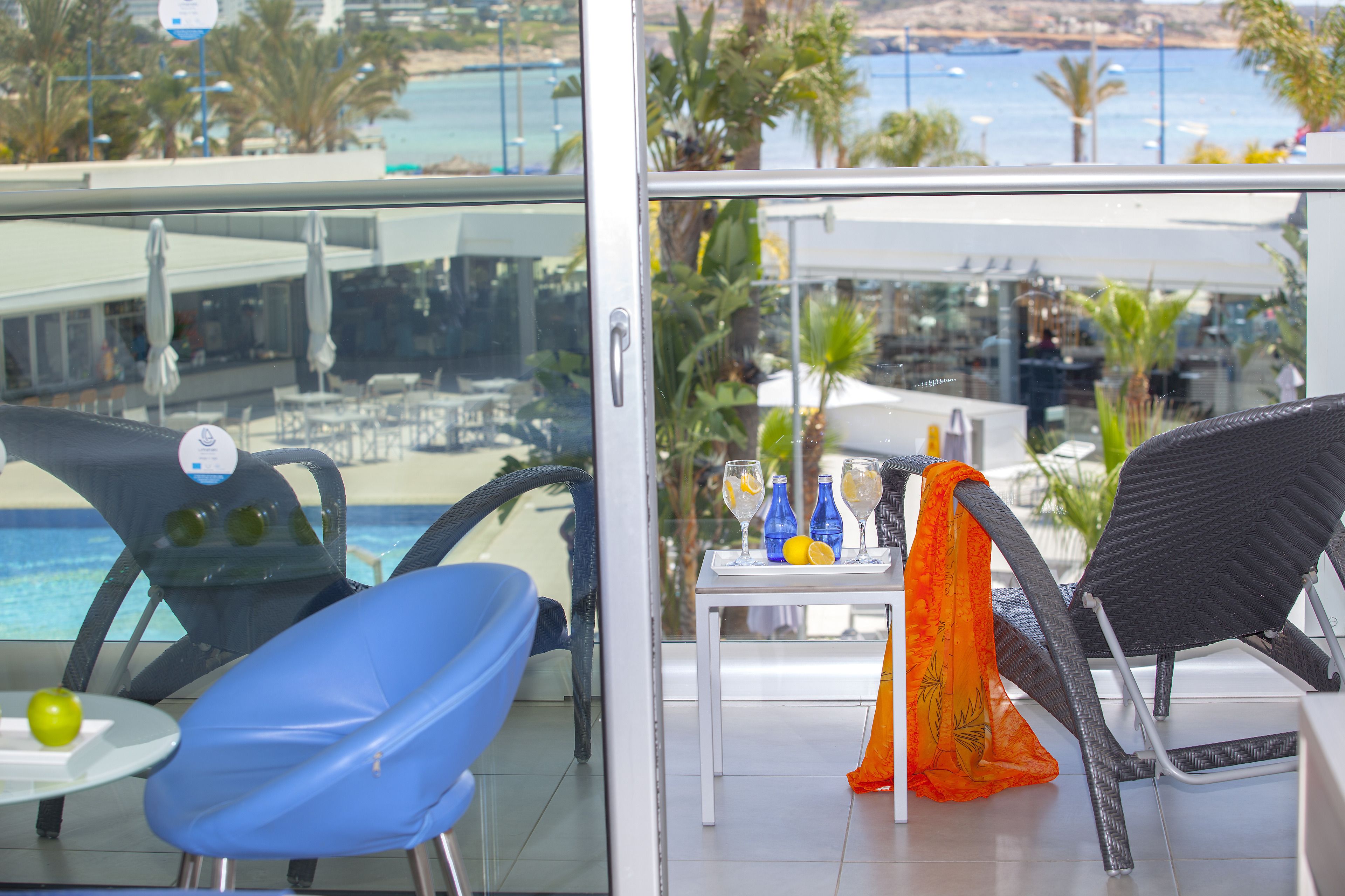 A balcony with a glass door, a table with drinks and a decorative orange cloth, and a view of the sea and palm trees.