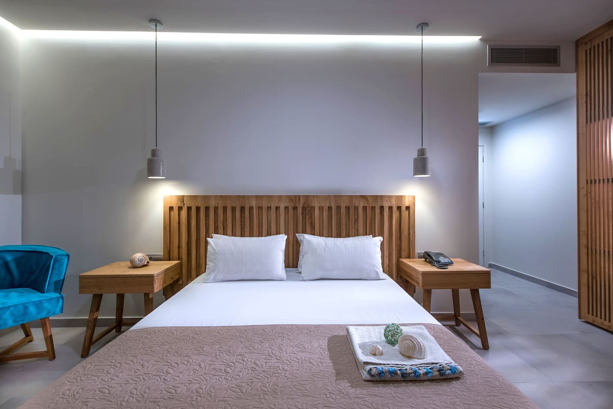 A modern hotel room with a wooden headboard, a large bed with white linens, two bedside tables with lamps, and a blue chair.