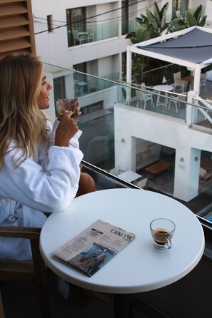 A woman in a white robe sitting at a round table with a newspaper and a glass of coffee. The table is on a balcony with a glass railing, overlooking a modern building with outdoor seating and plants.