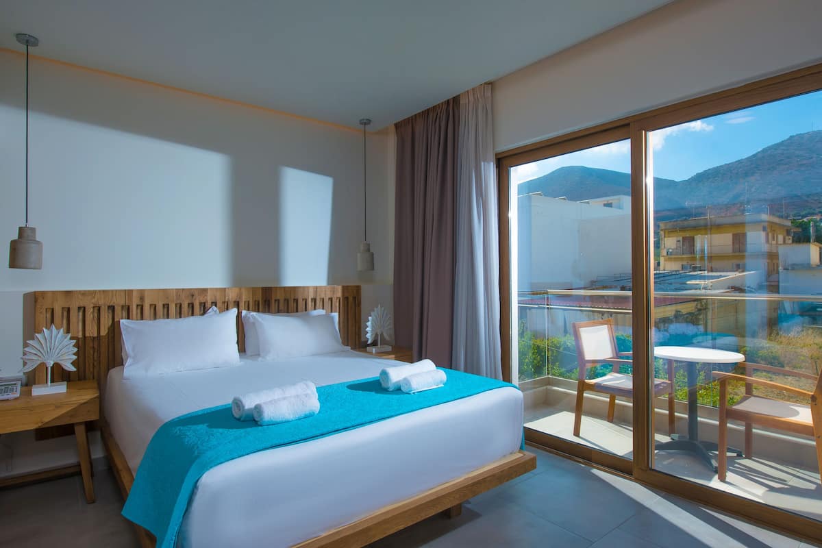 A hotel room with a large bed, a wooden headboard, a balcony with chairs, and a view of the mountains.