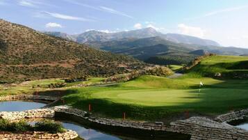 A golf course with a pond, surrounded by hills and mountains.