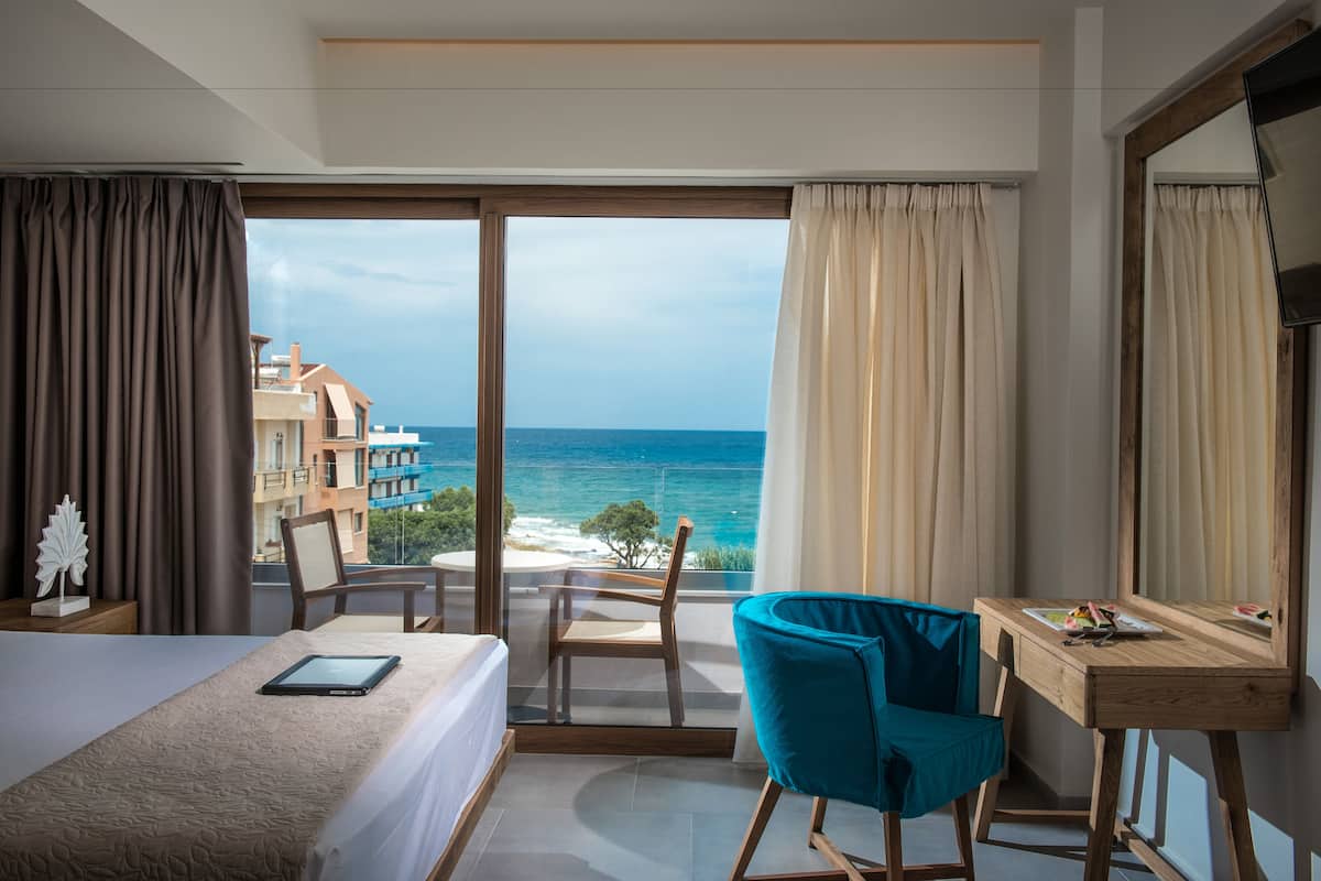 A hotel room with a bed, a desk, a chair, and a large window offering a view of the ocean and buildings.