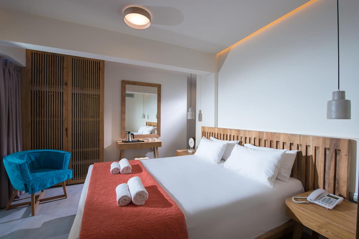 A hotel room with a large bed, a wooden headboard, a blue chair, a wooden wardrobe, a desk with a mirror, and a telephone on the bedside table.