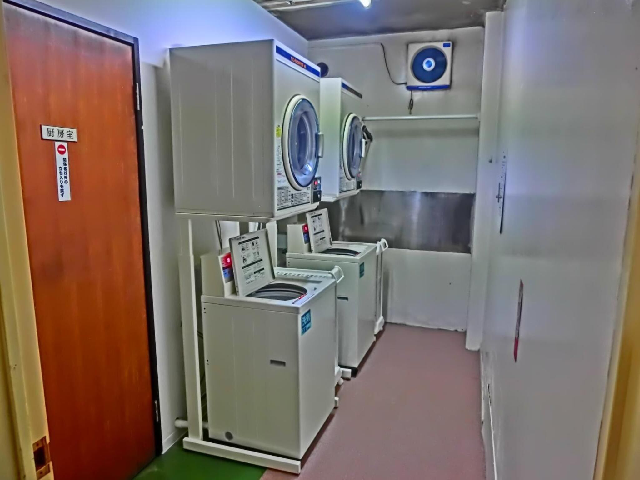 A laundry room with multiple washing machines and dryers, a wooden door, and a sign indicating it is a restricted area.