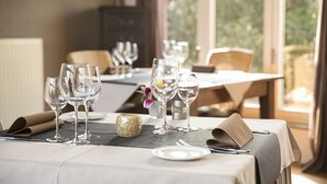 A dining table set with wine glasses, plates, and cutlery, with a view of greenery outside. - Hotel de Maretak (Dilsen-Stokkem)