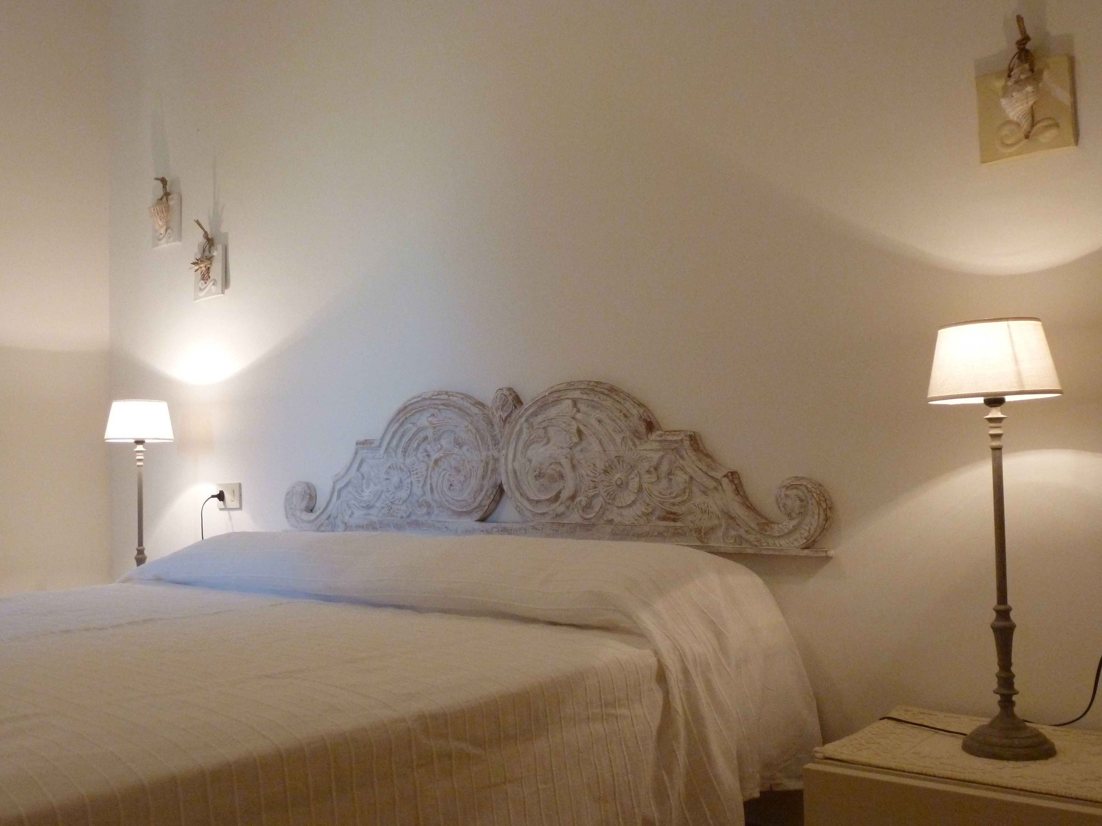 Cama bem arrumada com cabeceira branca, luminária de mesa e um quadro pendurado na parede.