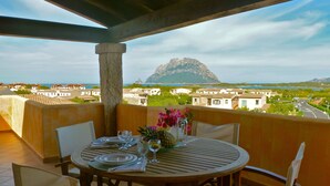 A round wooden table set for two with a view of a mountain and coastal town. - Il Borgo Appartamenti by Kasavacanze (Loiri Porto San Paolo)