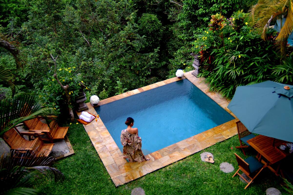 A rectangular pool with a tiled border, surrounded by lush greenery and a wooden deck with seating.