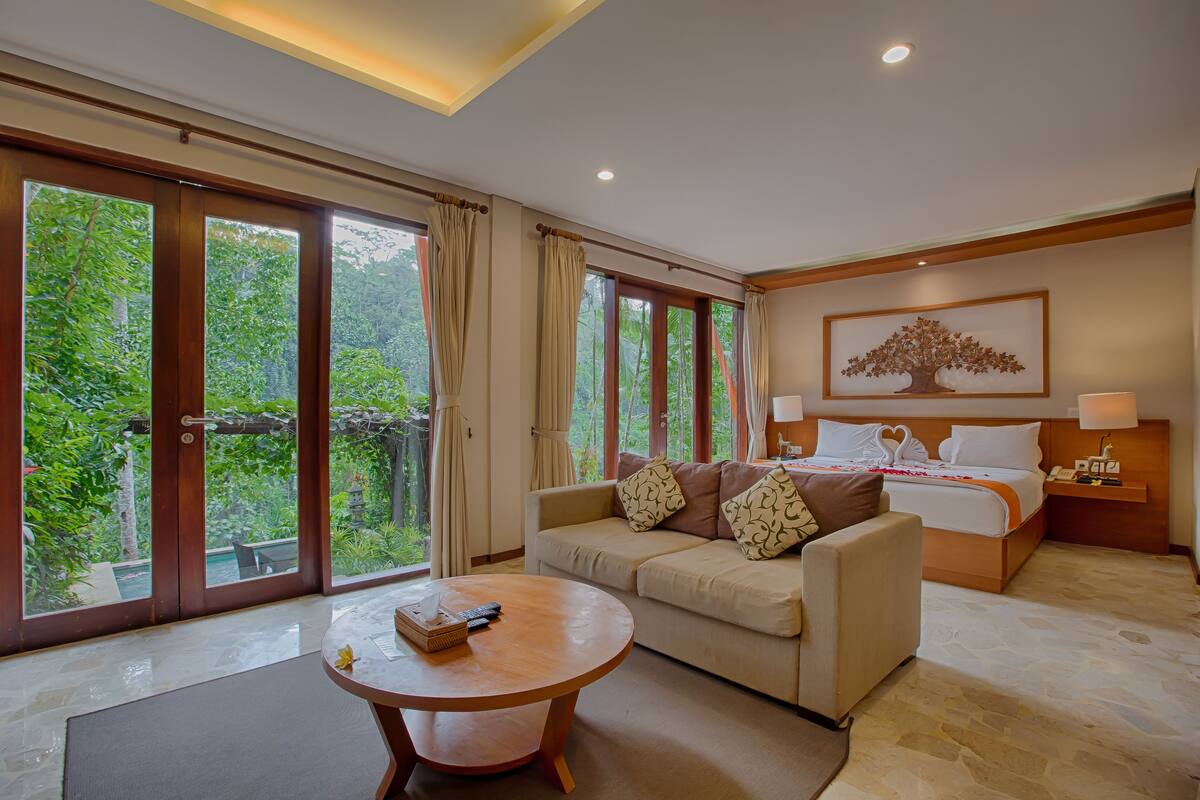 A hotel room with a bed, a sofa, a coffee table, and large windows offering a view of greenery.