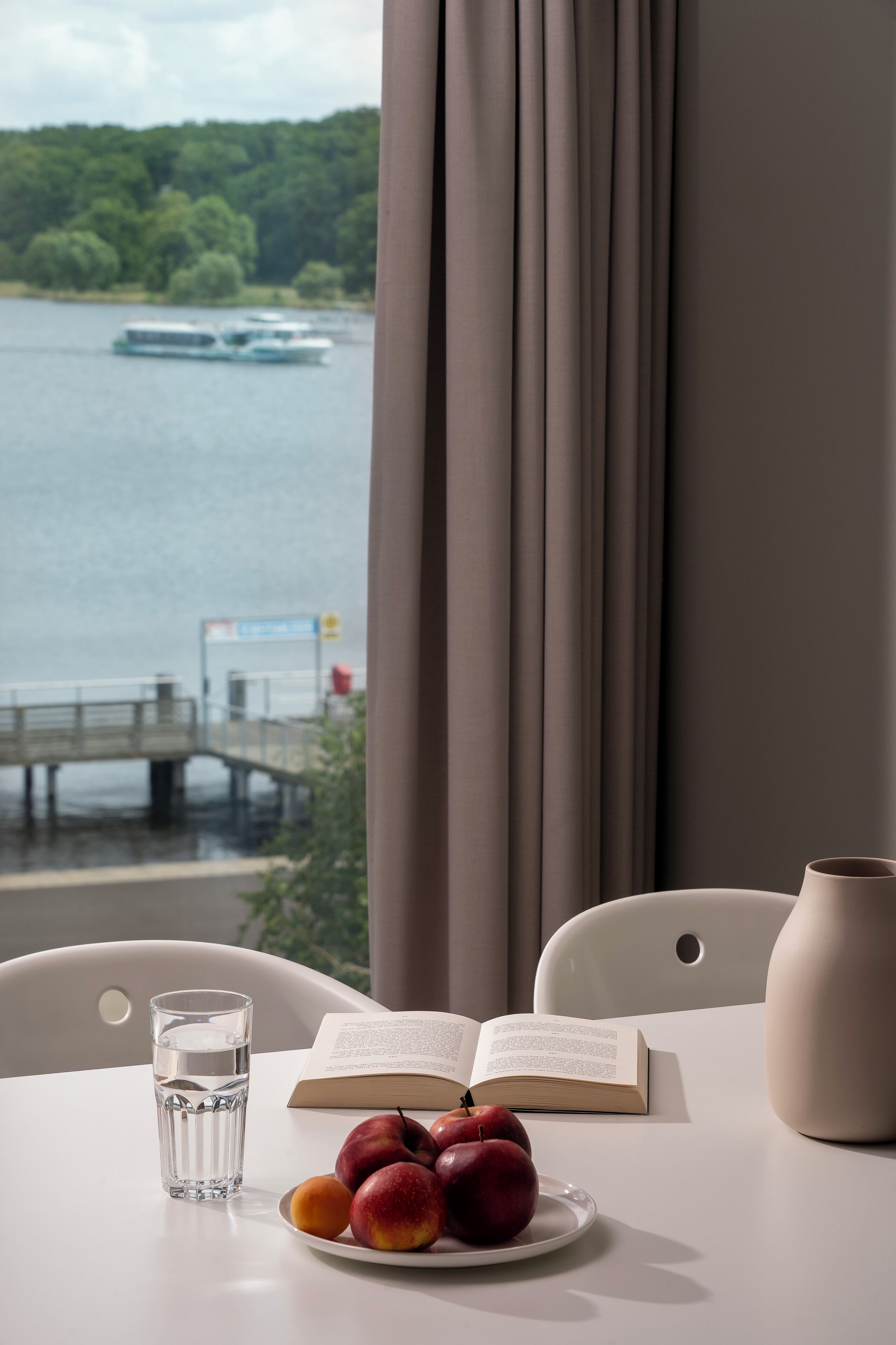 Une table avec un verre d’eau, un livre ouvert et une assiette de fruits, placée près d’une fenêtre donnant sur un lac et une jetée.
