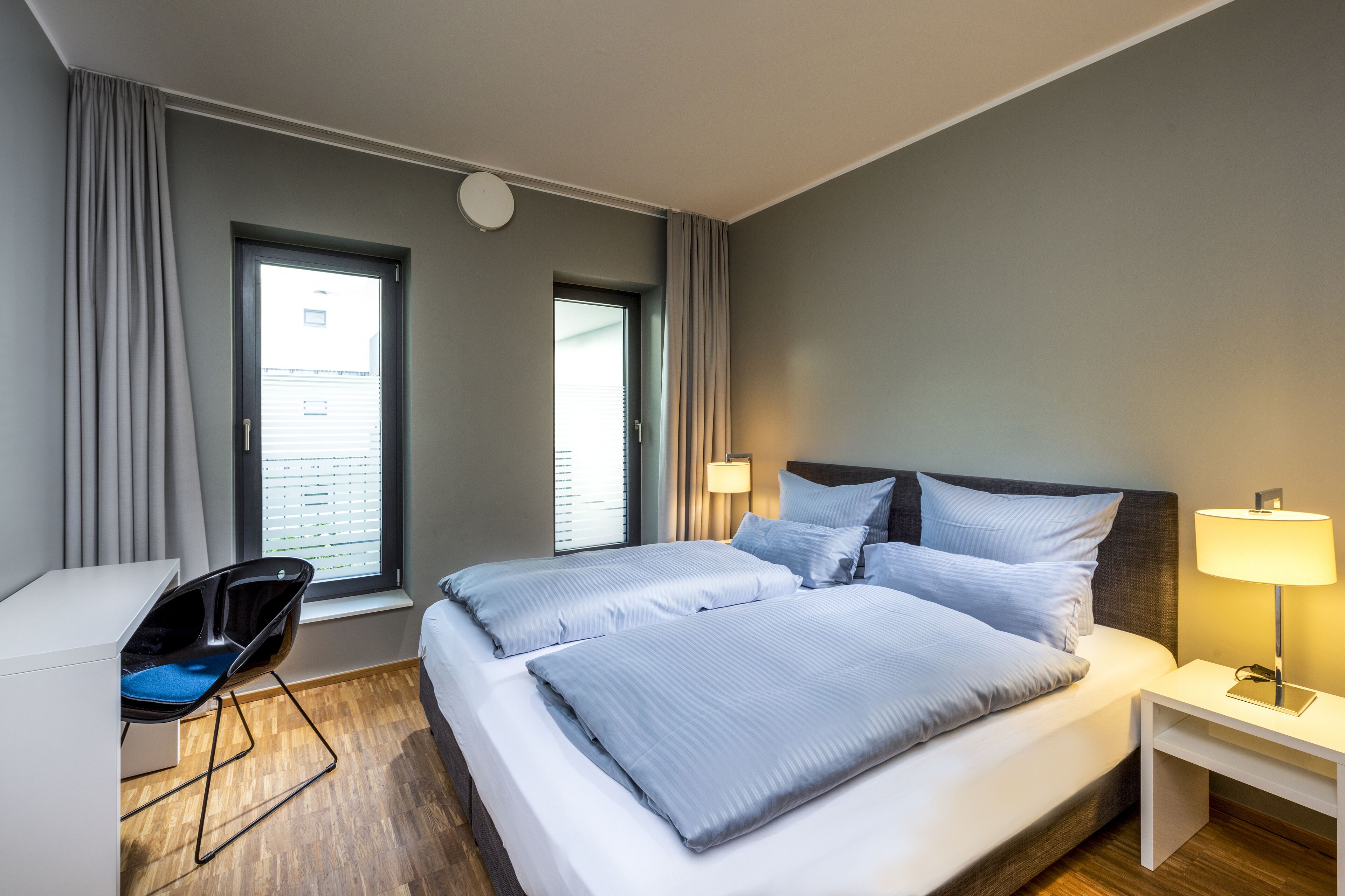 Une chambre d’hôtel moderne avec un grand lit, un bureau et une chaise. Elle comprend deux fenêtres avec des rideaux, une table de chevet avec une lampe et un luminaire au plafond.