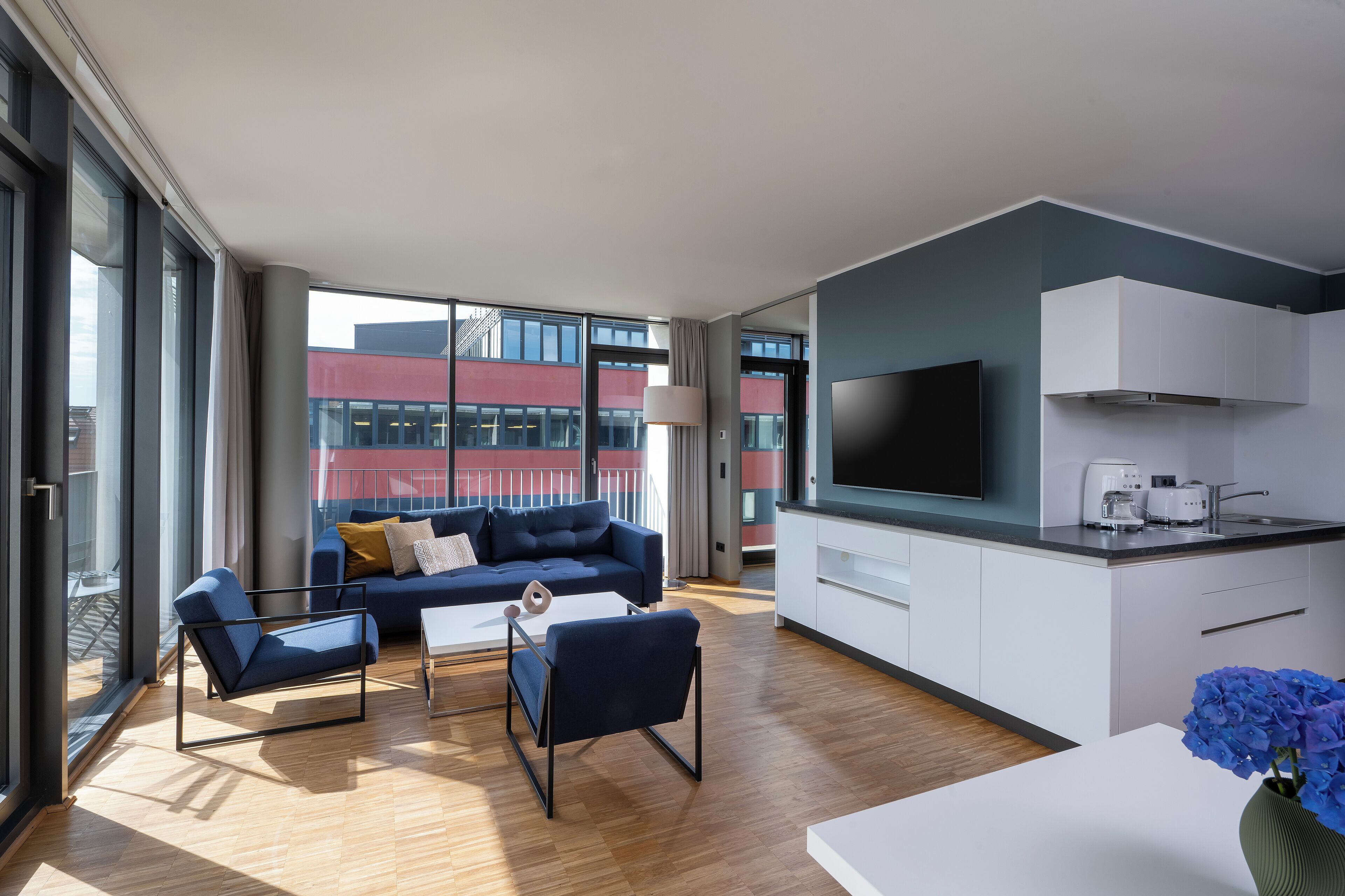 A modern living room with a sofa, chairs, and a coffee table. There is a kitchen area with a TV, cabinets, and a window.