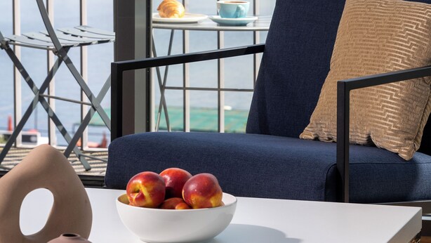 A balcony with a view of the sea, a table with a bowl of fruit, a chair, and a small table with a cup and pastry. - Waveboard Hotel & Boardinghouse  (Potsdam)