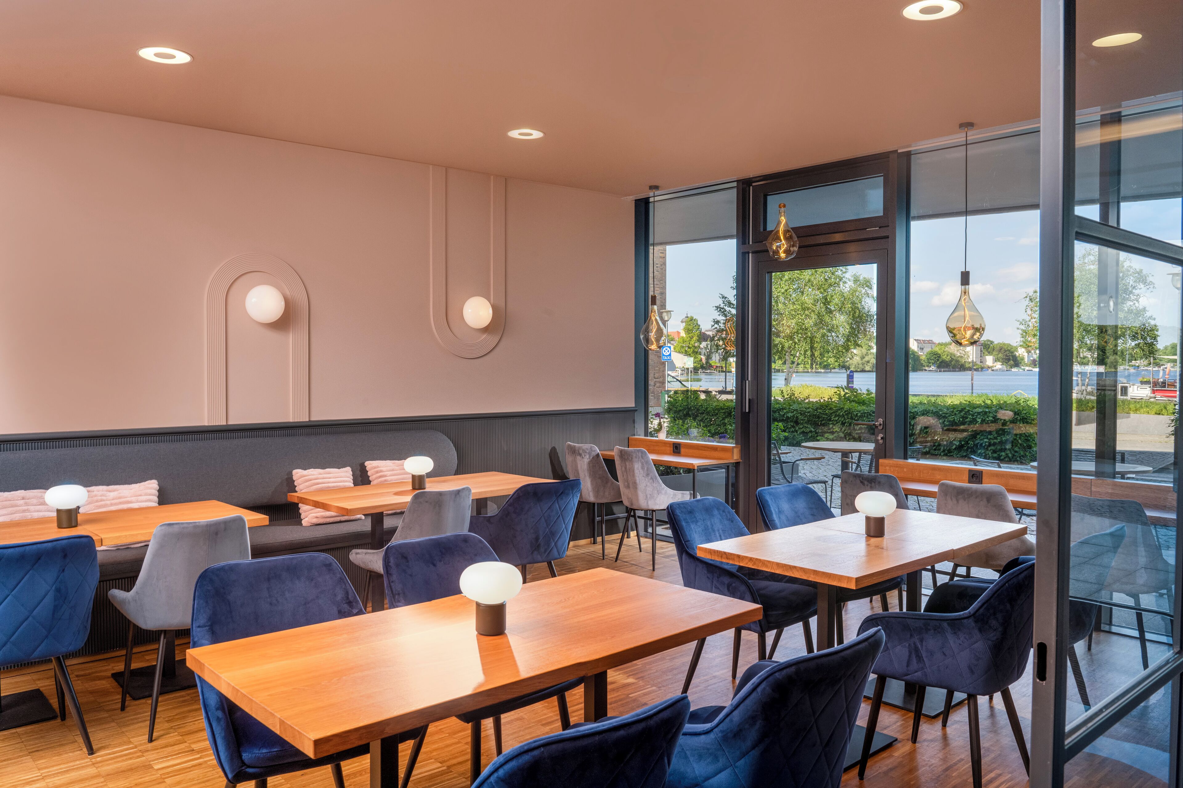 Un restaurant moderne avec des tables en bois et des chaises bleues, de grandes fenêtres donnant sur l’extérieur et des luminaires suspendus.
