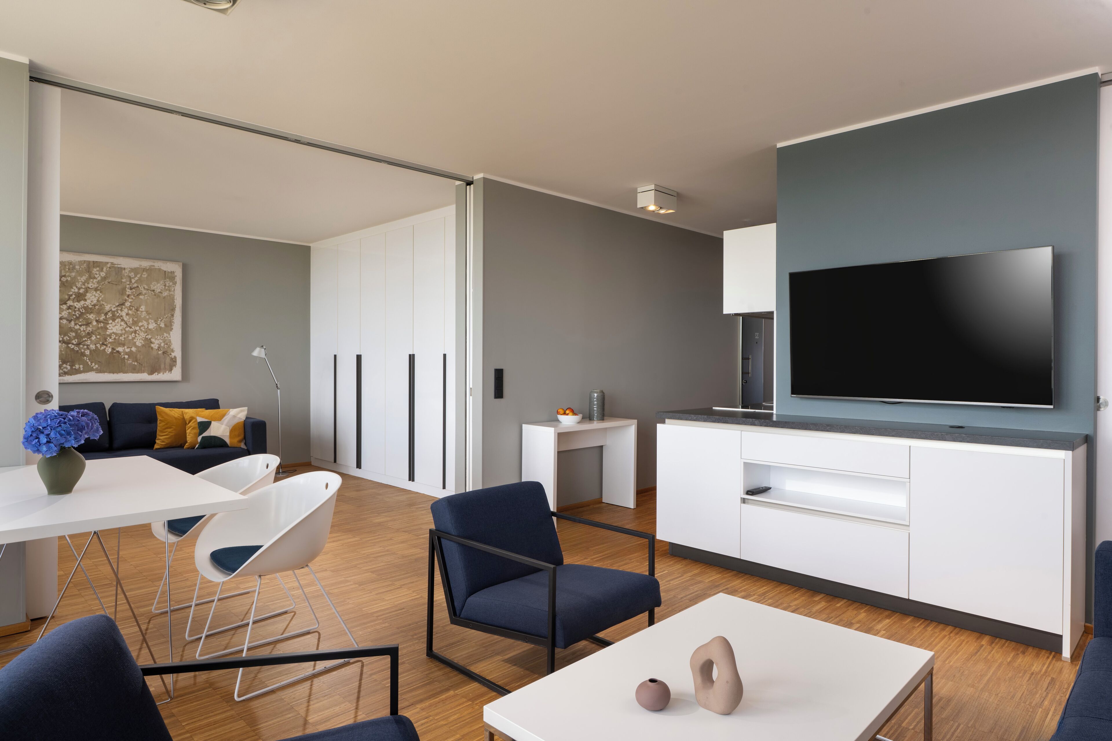A modern living room with a flat-screen TV, white cabinetry, a dining area with a white table and chairs, and a sofa with cushions.