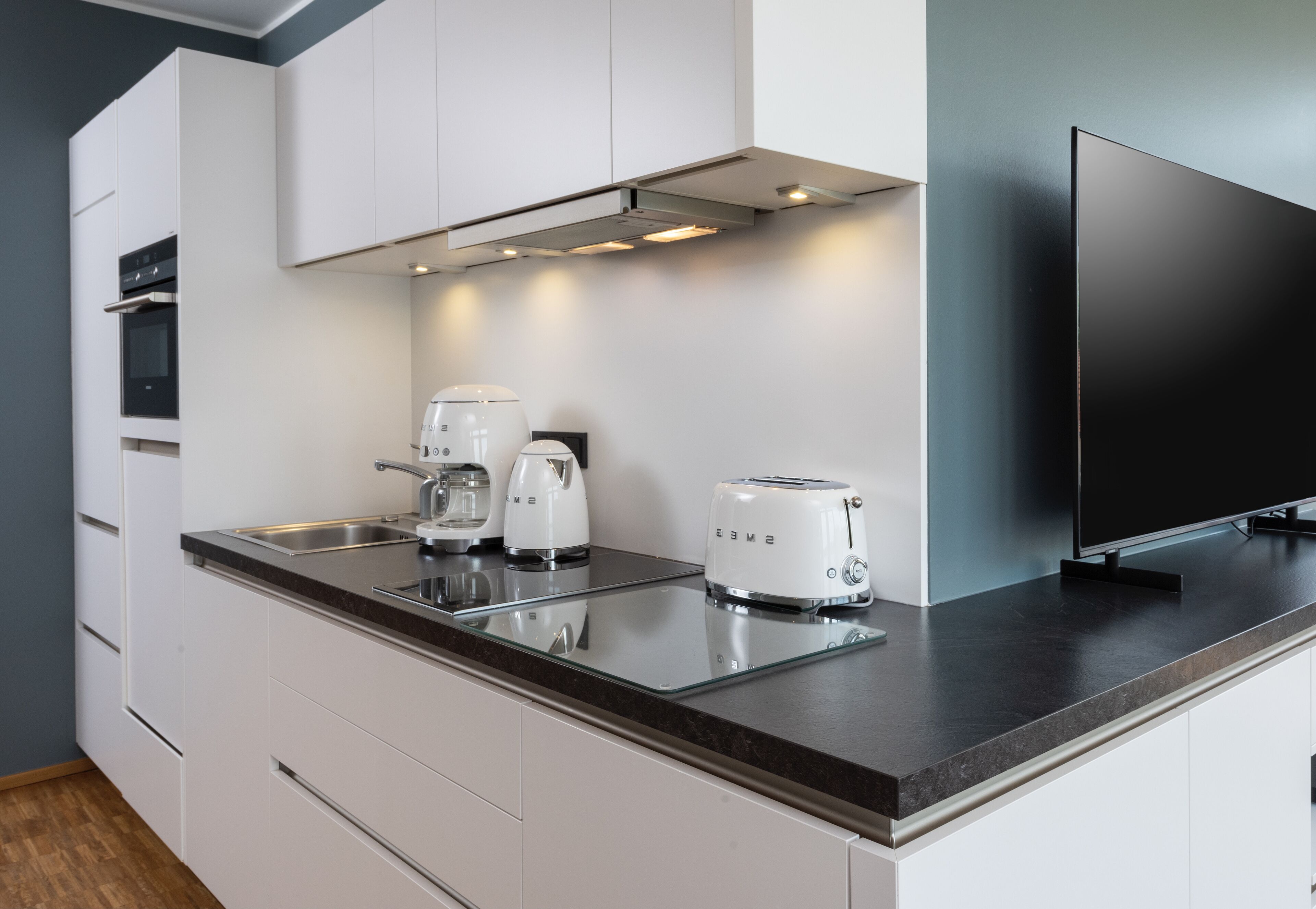 Eine moderne Küche mit weißen Schränken, einer schwarzen Arbeitsplatte, einer Kaffeemaschine, einem Toaster und einem Flachbildfernseher.