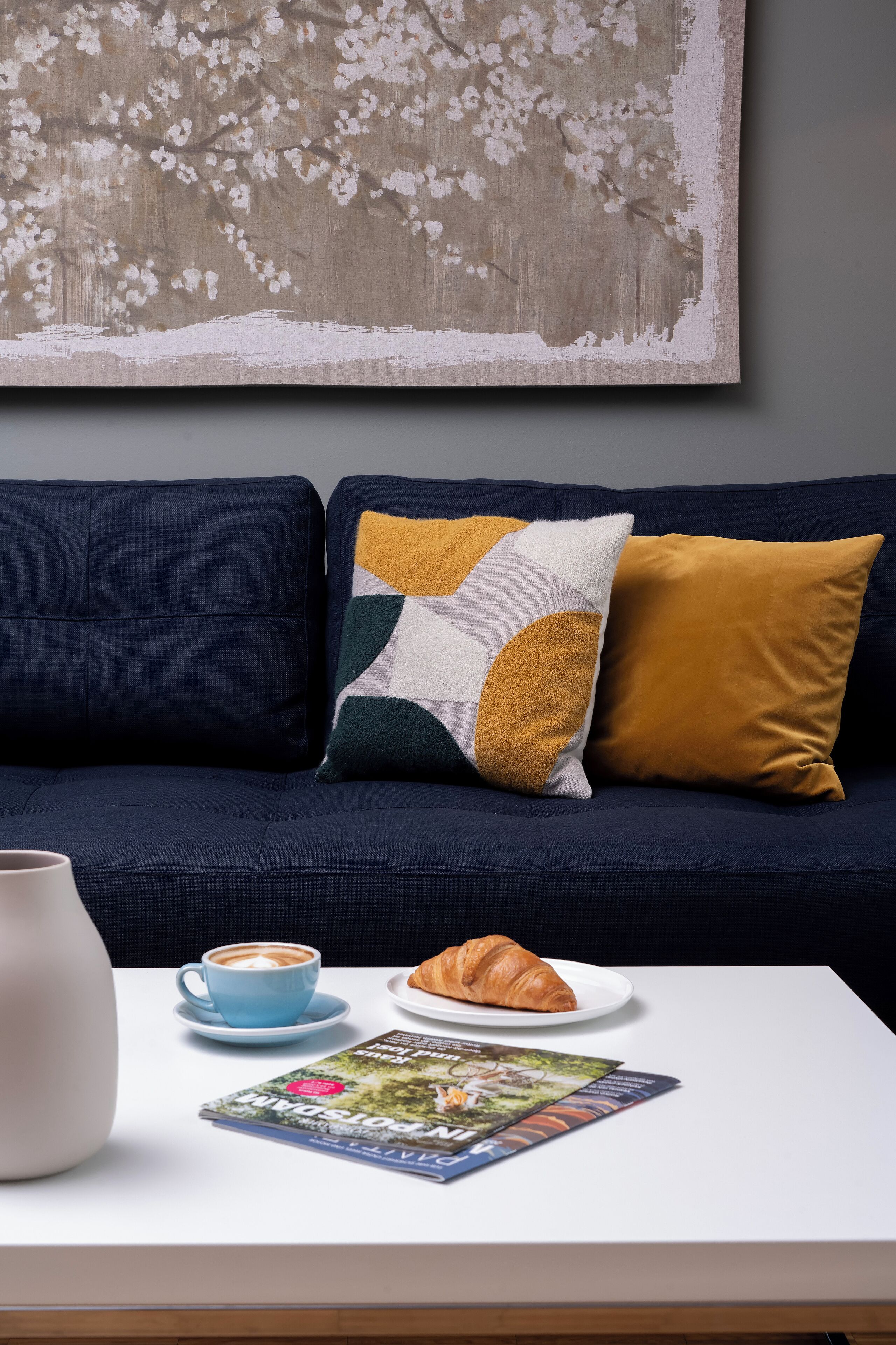 Een donkerblauwe bank met twee kussens, een witte vaas, een blauw kopje met een latte, een croissant en een tijdschrift op een witte tafel.