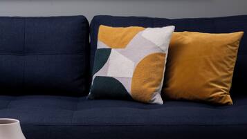 Een donkerblauwe bank met twee kussens, een witte vaas, een blauw kopje met een latte, een croissant en een tijdschrift op een witte tafel.