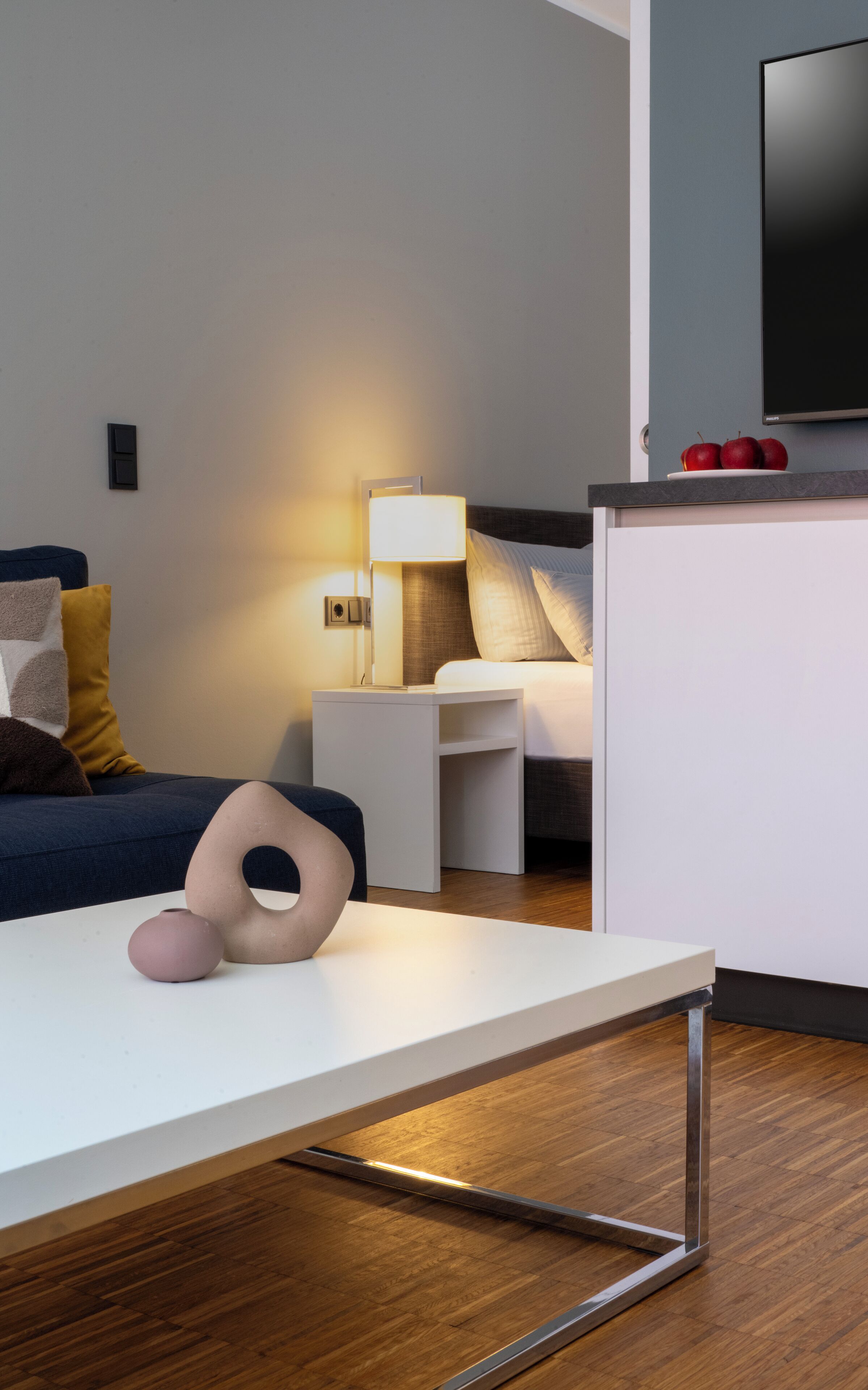 A modern hotel room with a bed, bedside table, lamp, and a flat-screen TV on a stand.