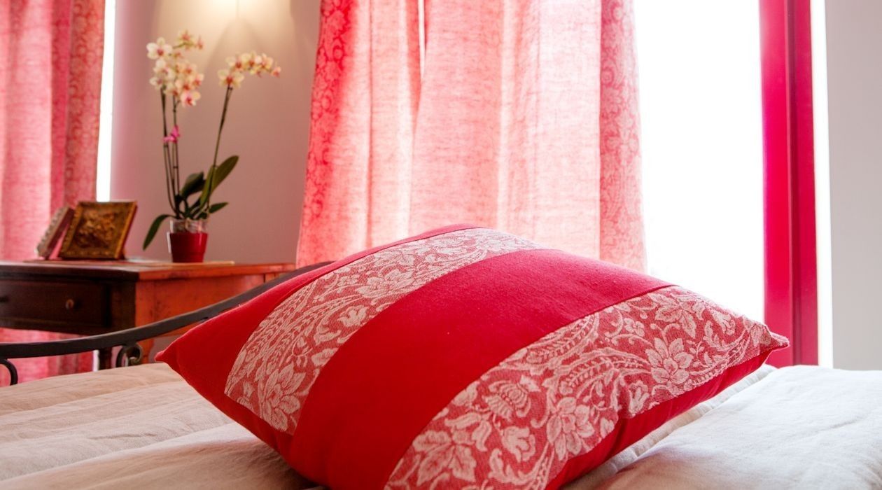A bed with a red and white pillow, a wooden nightstand with a lamp, and a window with pink curtains and an orchid plant.