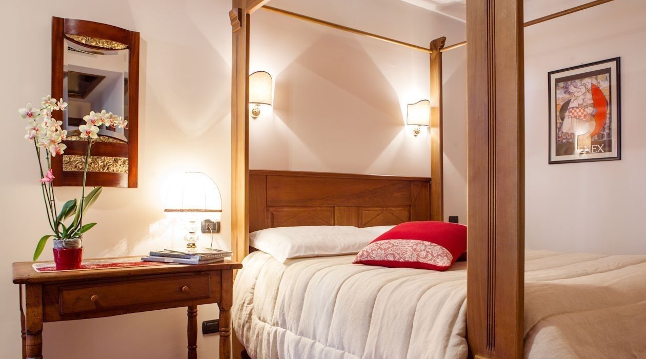 Un letto a baldacchino con biancheria bianca e un cuscino rosso, un comodino in legno con una lampada, uno specchio e un'opera d'arte incorniciata al muro.