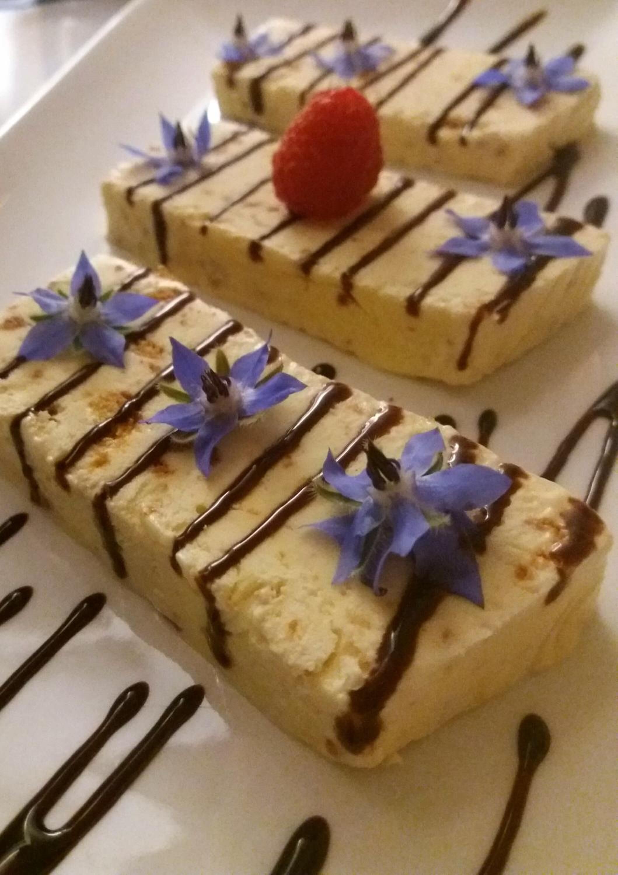 A dessert plate with rectangular pieces of cake, drizzled with chocolate sauce and garnished with blue flowers and a strawberry.