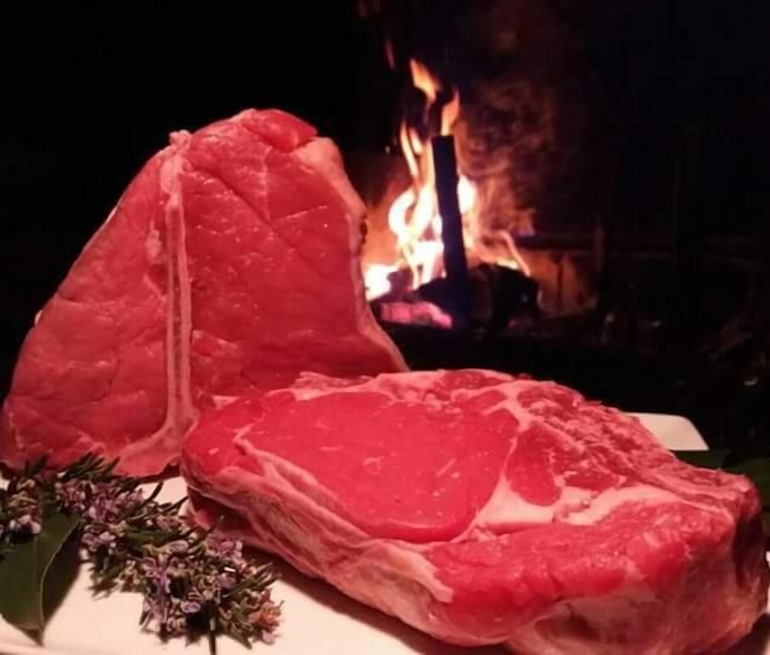 Ein rohes Steak auf einem weißen Teller, daneben ein Blumenstrauß, vor dem Hintergrund eines brennenden Kamins.