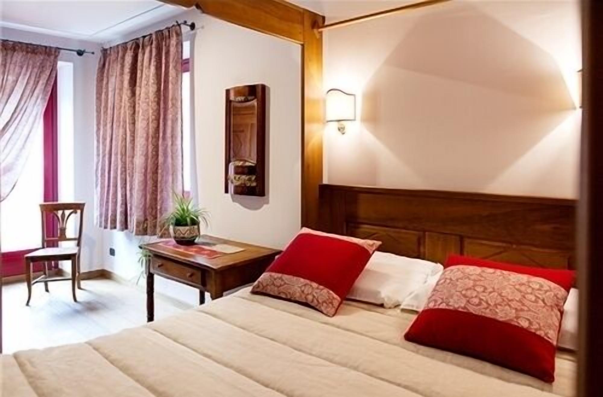 Una habitación de hotel con cama, dos almohadas, una mesita de noche, una silla, un escritorio, una lámpara y una ventana con cortinas.