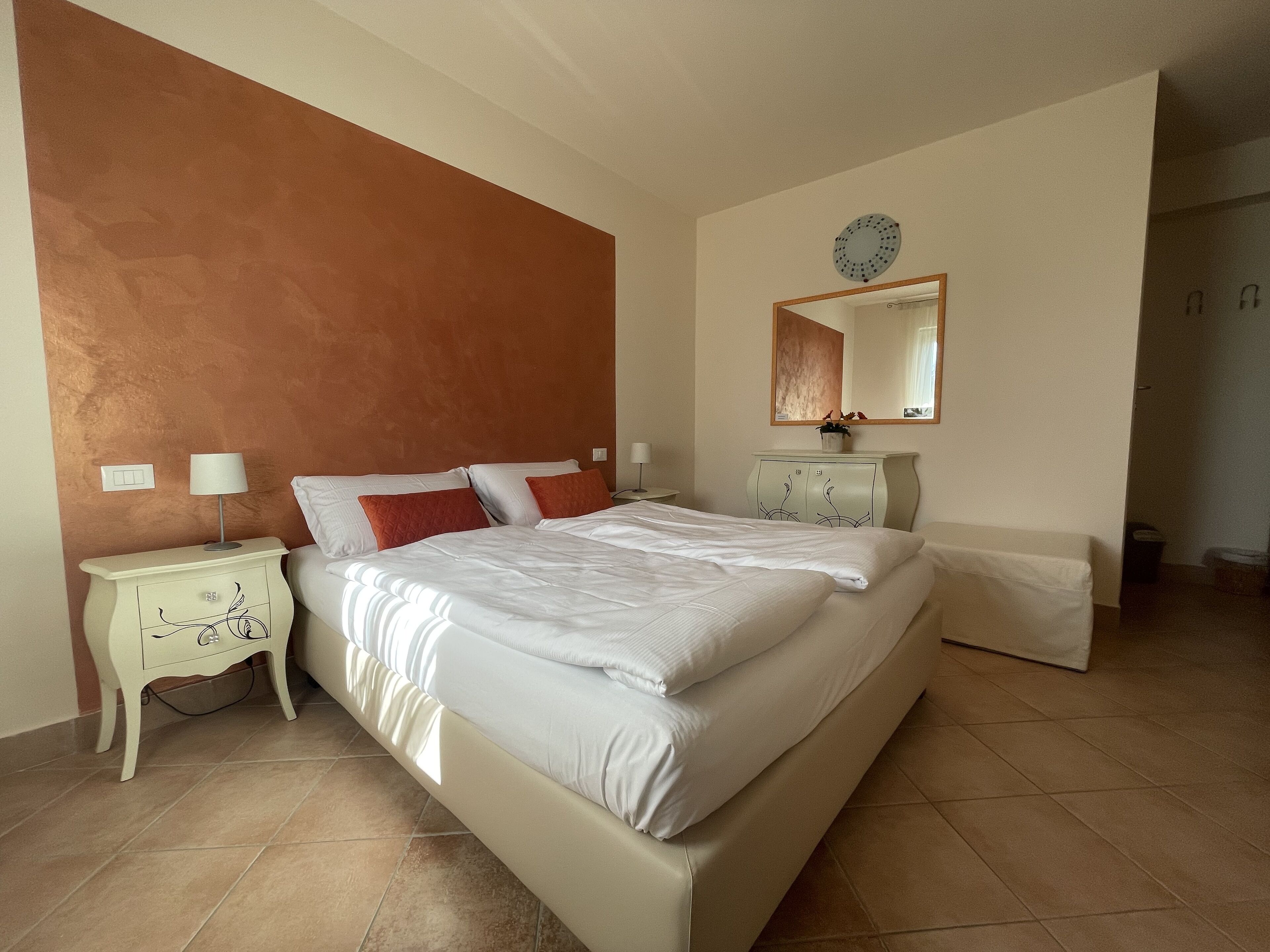 Un dormitorio con una cama grande, una mesita de noche con lámpara, un aparador y un reloj de pared.