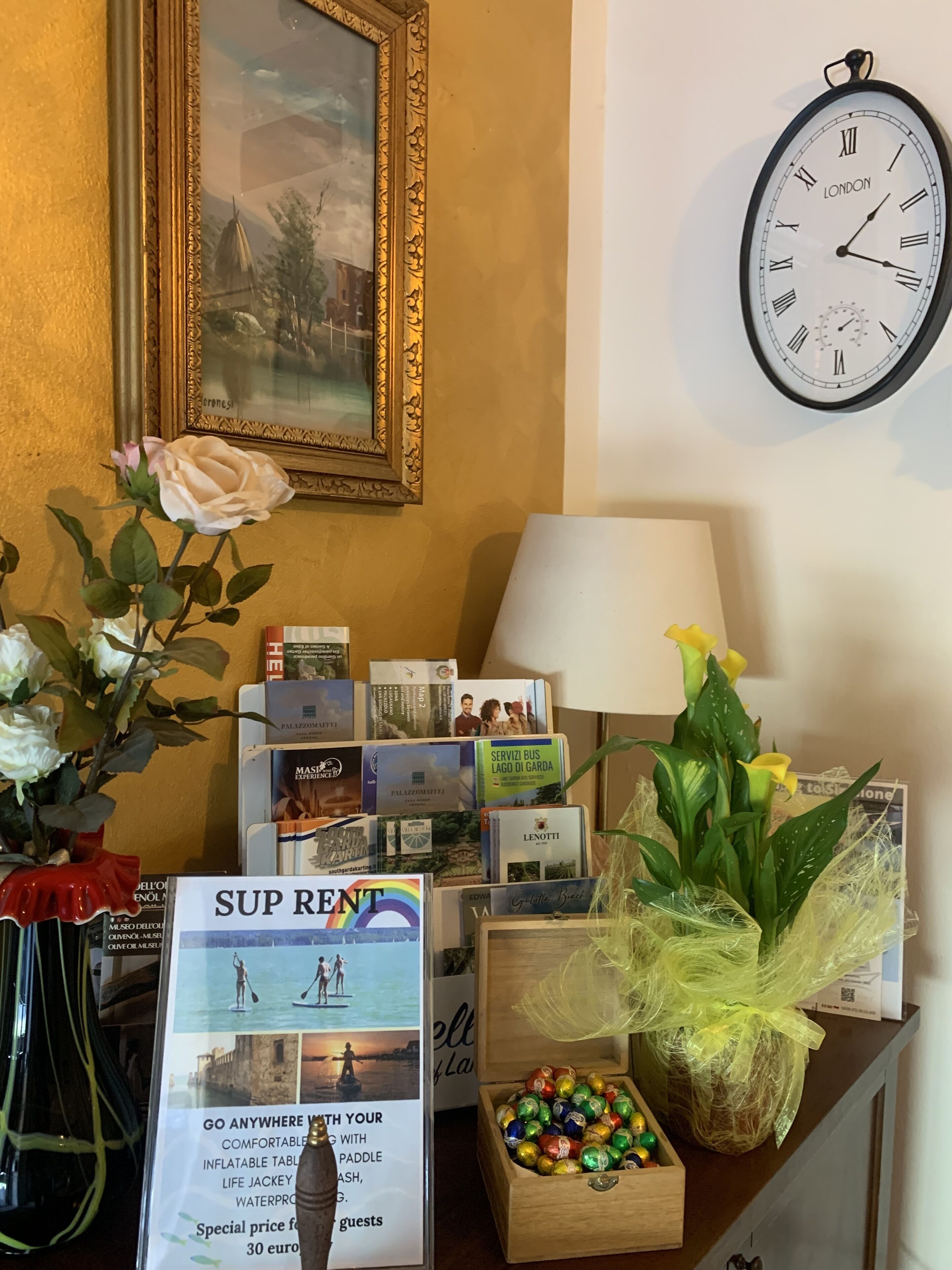 Ein Schreibtisch mit einem gerahmten Gemälde, einer Uhr, einer Vase mit Blumen, einer Lampe, einem Schild und einer Schachtel Pralinen.