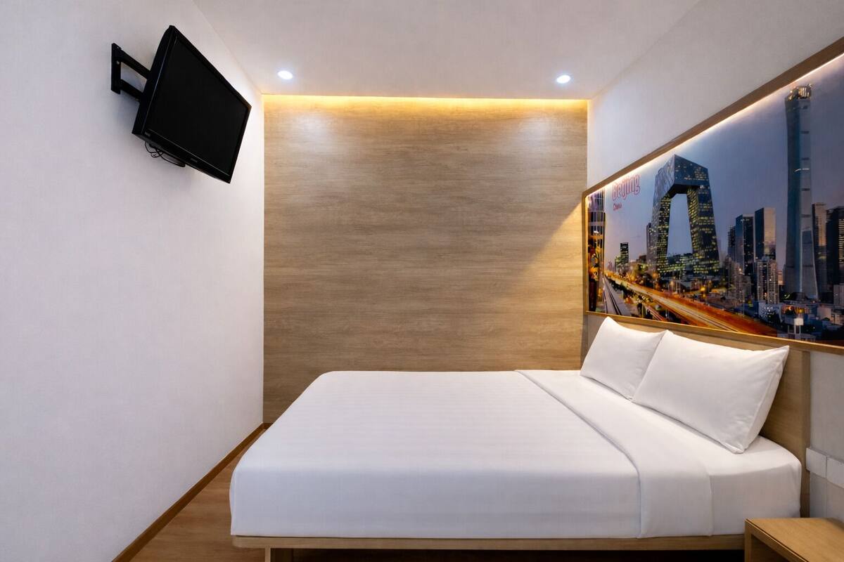 A modern hotel room with a bed, a mounted TV, a cityscape painting, and a wooden accent wall.
