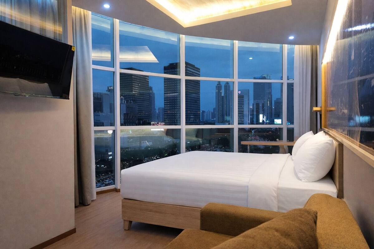 A hotel room with a large window offering a city view at night, a bed, a TV mounted on the wall, and a small table.