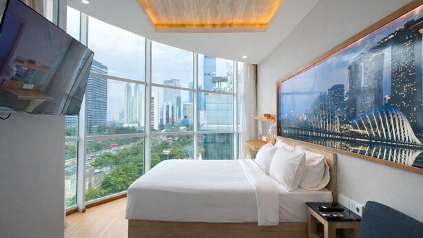 A modern hotel room with a large bed, a TV mounted on the wall, and a cityscape view through large windows.