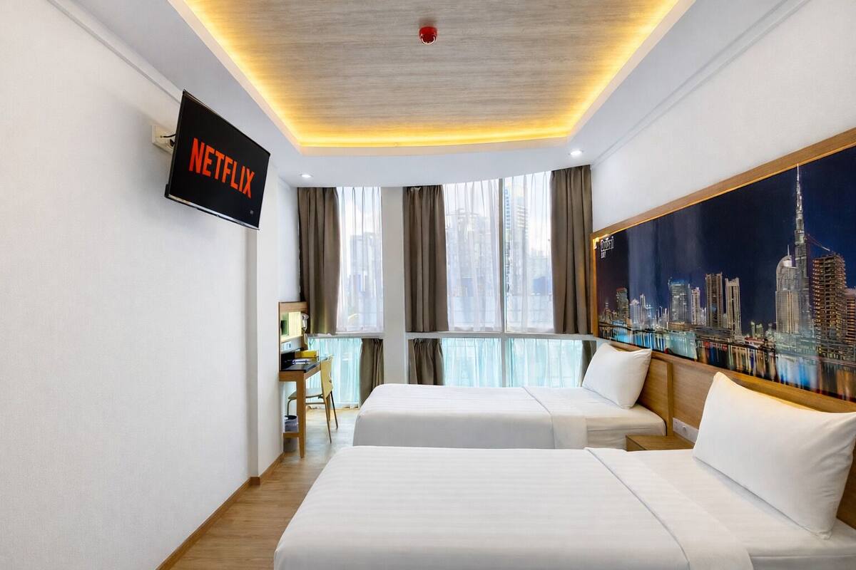 A hotel room with a bed, a desk, a chair, a TV mounted on the wall, and a large window with curtains.