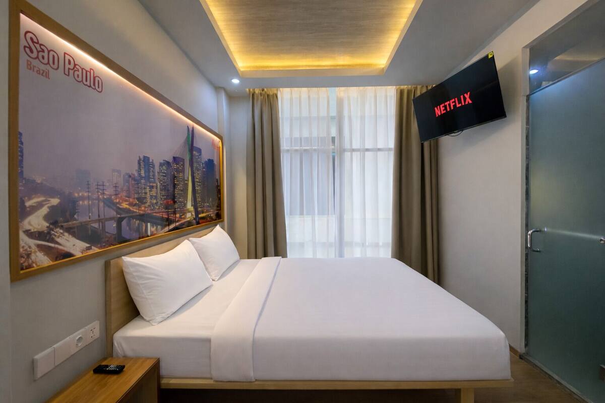 A hotel room with a bed, a TV mounted on the wall, a poster of São Paulo, and a window with curtains.
