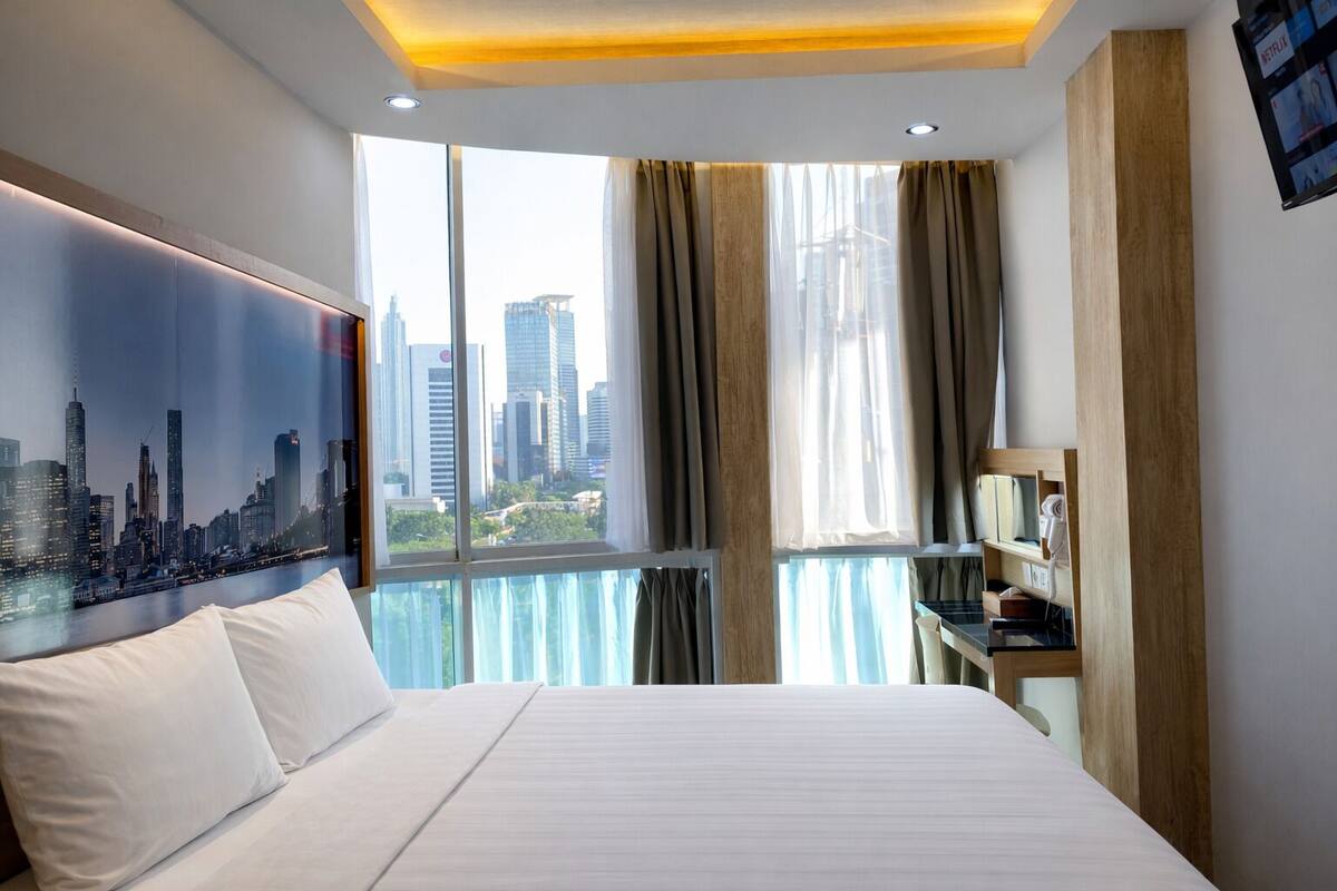 A modern hotel room with a large bed, a cityscape view, a desk with a lamp, and a mounted TV displaying Netflix.