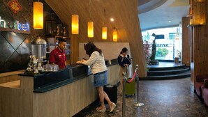 A hotel reception area with a wooden reception desk, a coffee machine, and a statue. There are hanging pendant lights and a statue in the background.