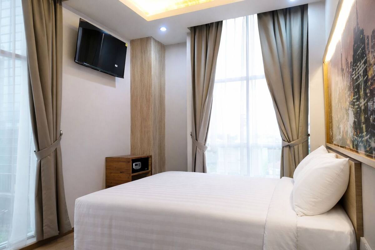 A hotel room with a bed, a TV mounted on the wall, a nightstand, and a window with curtains.