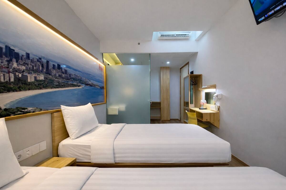 A hotel room with a bed, a desk with a chair, a mirror, and a painting of a beach scene.