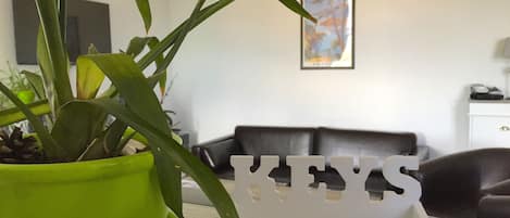 Una sala de estar con una planta verde en maceta, un adorno blanco con letras, un sofá de cuero negro y un cuadro en la pared.