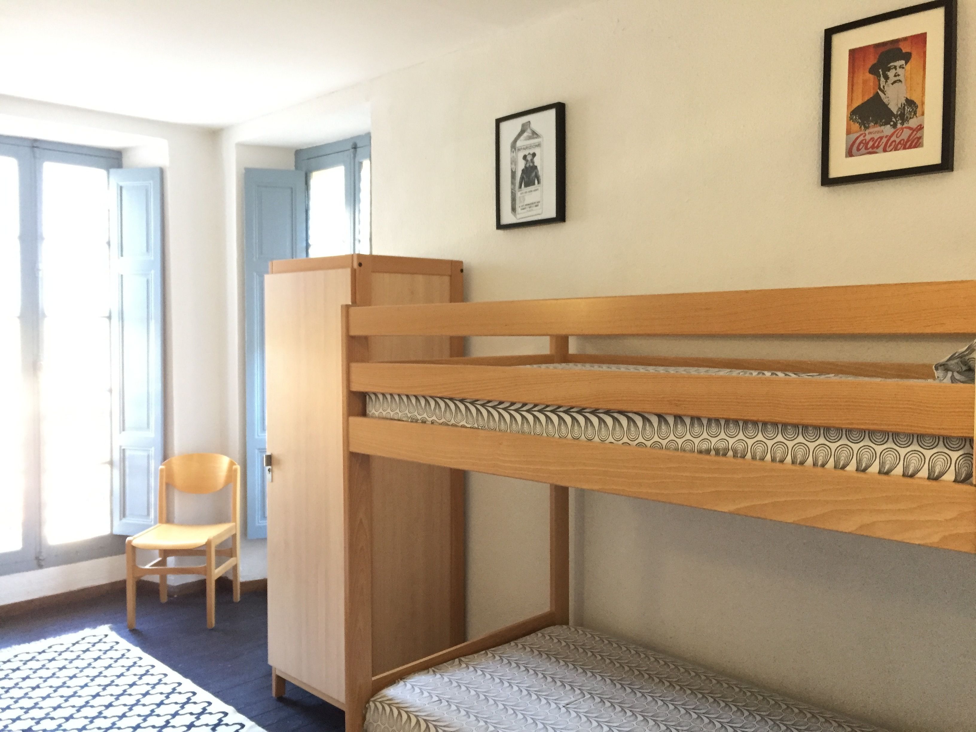 A room with a bunk bed, a wooden wardrobe, a chair, and two framed pictures on the wall.