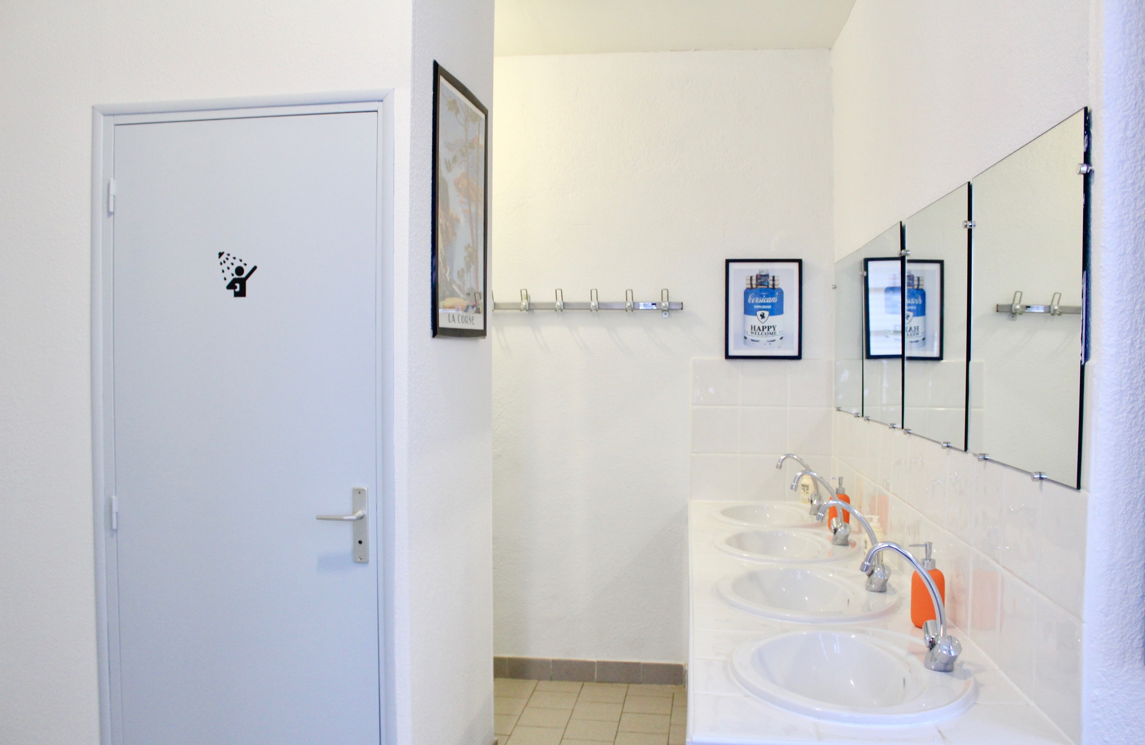 Une salle de bain avec une porte blanche, un distributeur de gel hydroalcoolique et un miroir au-dessus de deux lavabos.