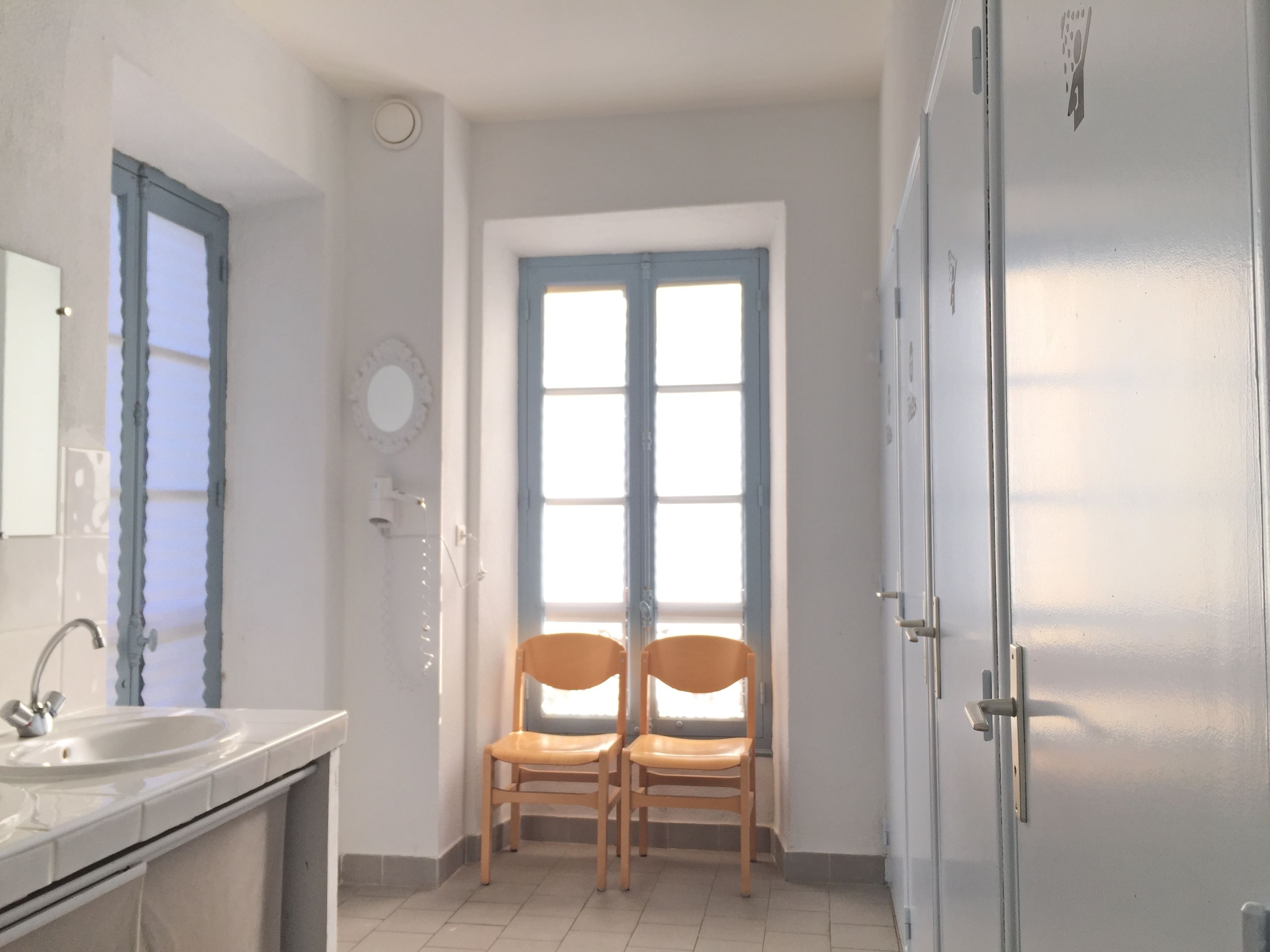Ein Badezimmer mit Waschbecken, Spiegel, zwei Holzstühlen und einem Fenster mit blauen Rollläden.
